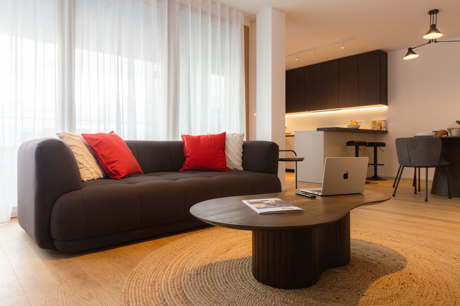 Lounge area with a textile comfortable sofa with plush cushions, a coffee table with a laptop on it.
On the left side is the tall kitchen countertop with tall chairs