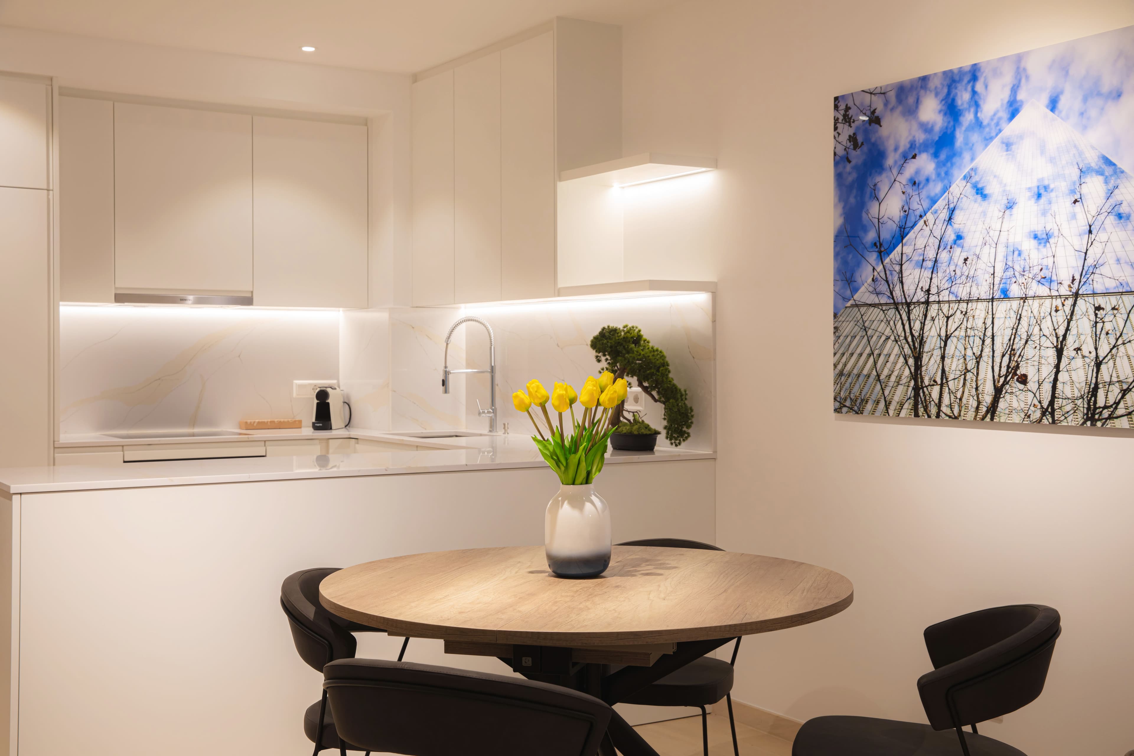 Round table with four chairs and yellow tulips, kitchen counter with a bonsai, and a coffee machine in the background. Louvre picture on the right wall.