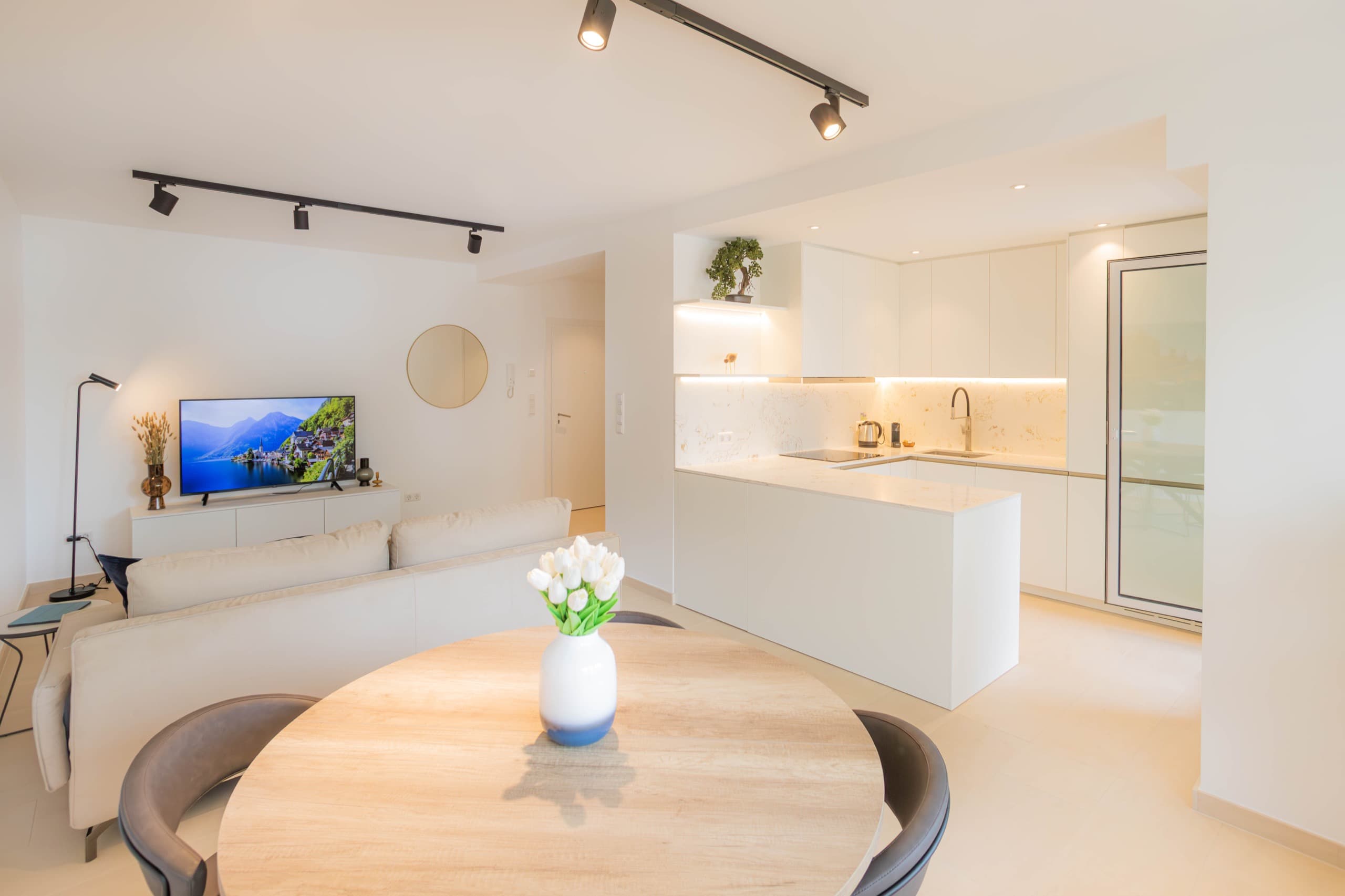 Open plan lounge with dining table and chairs, a sofa, TV stand with a smart TV on it. An oval mirror is in the hallway. Fully equipped kitchen is on the right.