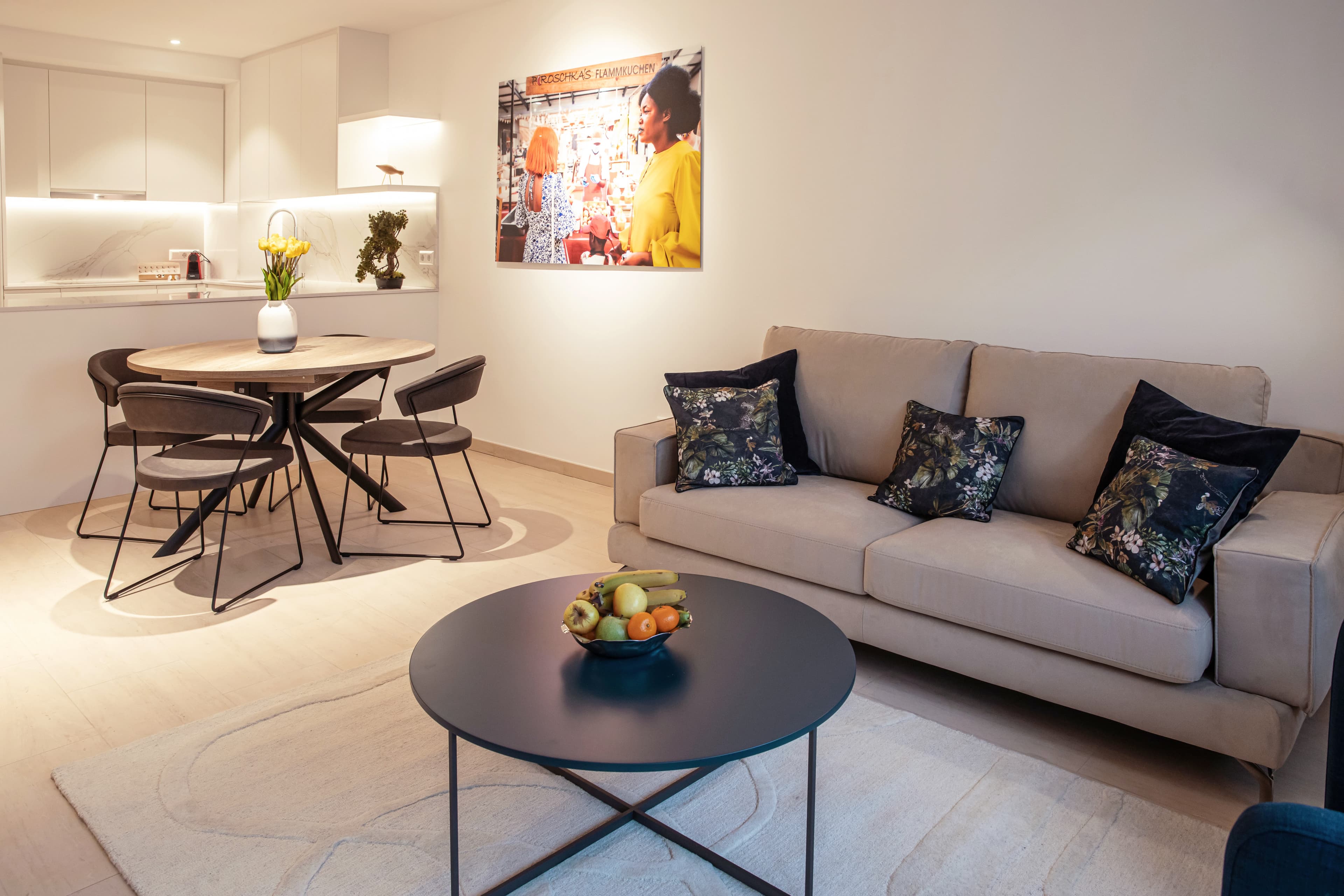 Lounge area with round dining room table and chairs, and a flower vase on top. Behind, there is a couch with a coffee table with a fruit bowl on it. Underneath the coffee table, there is a white carpet.