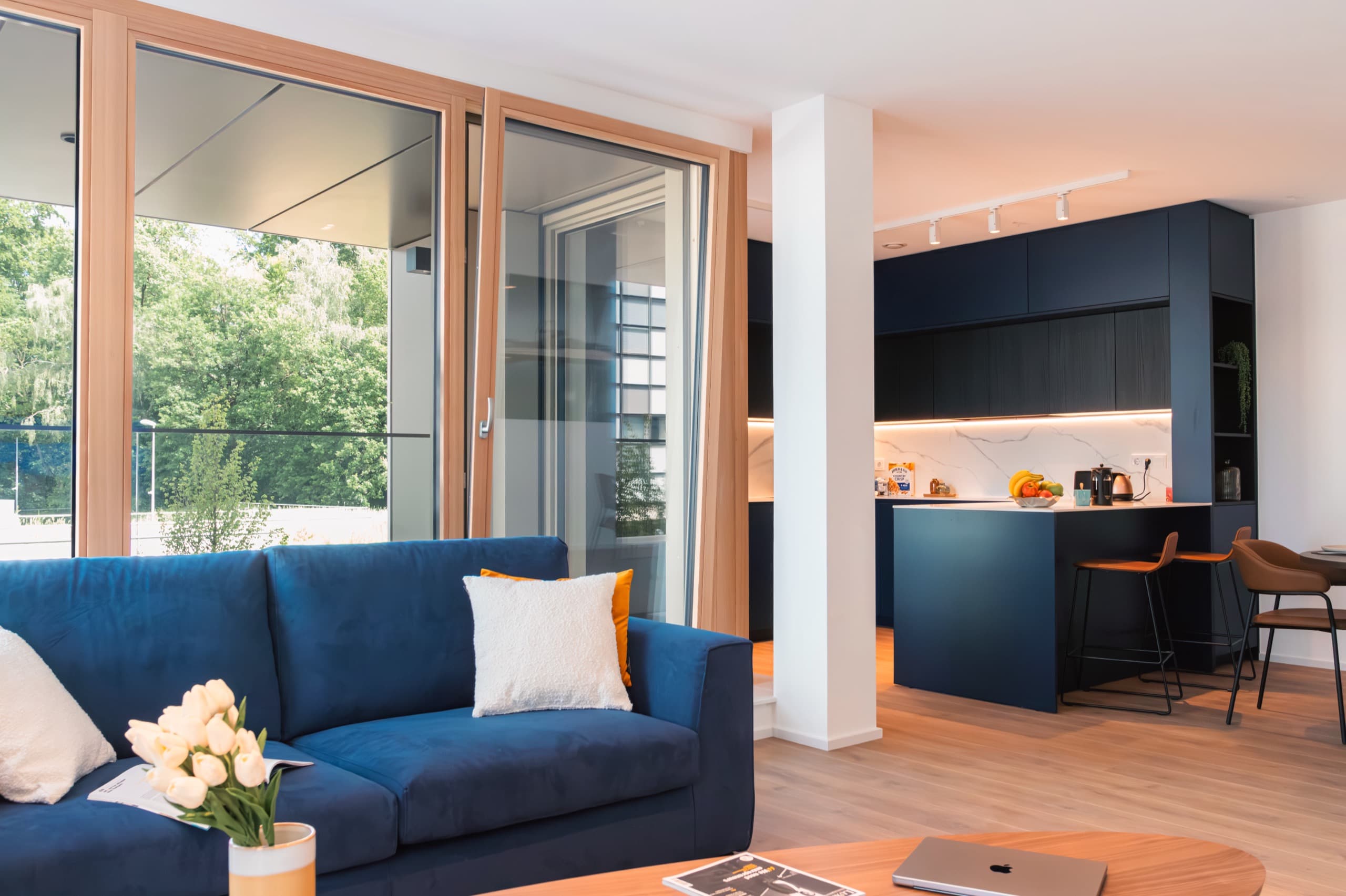 Overview of kitchen and lounge. Blue sofa with plush cushions, floor-to-ceiling windows behind. Tall kitchen countertop and a fully equipped kitchen. View over the forest behind the apartment's building.